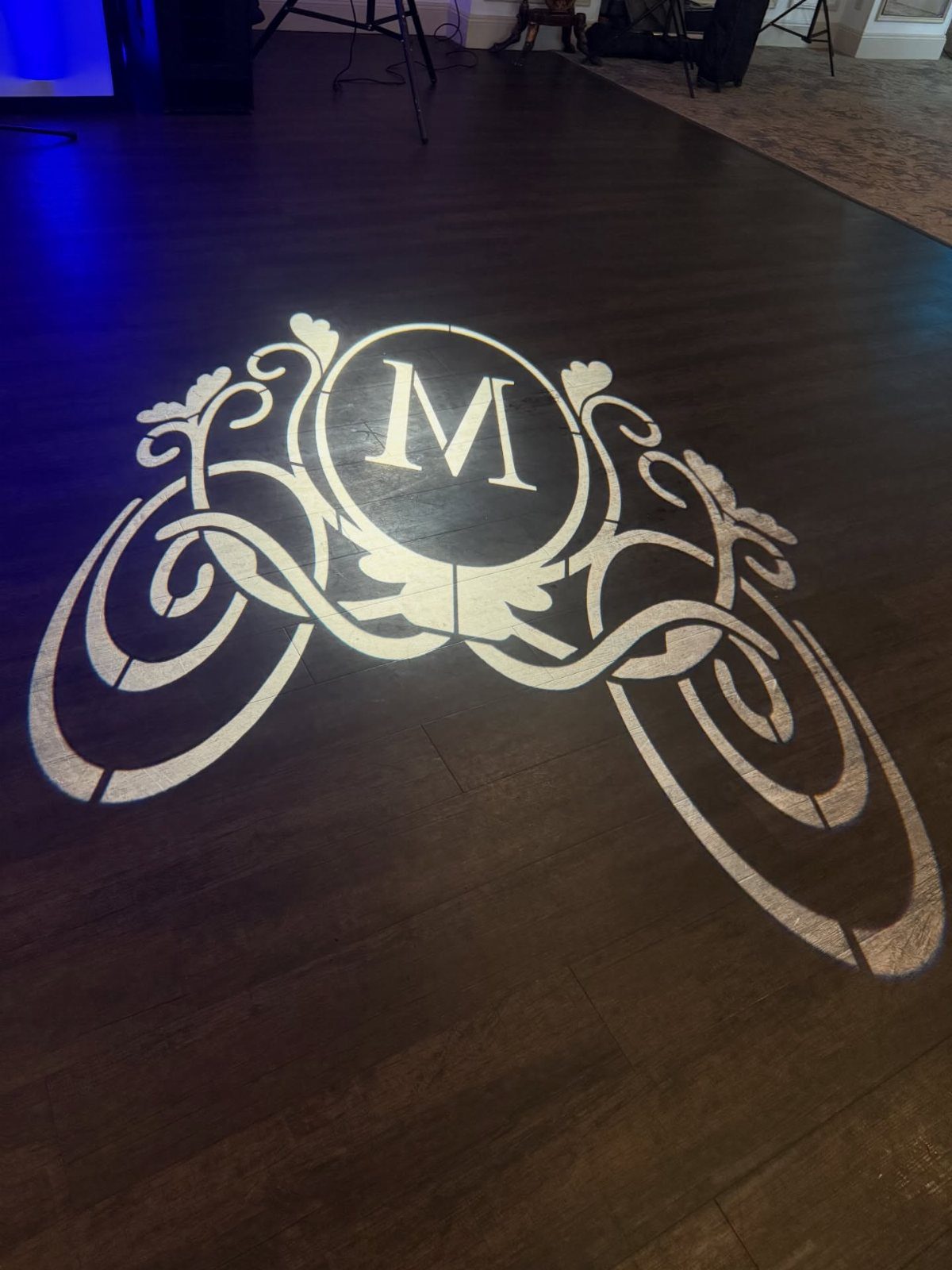 White monogram projection of the letter M on a dark wooden dance floor.