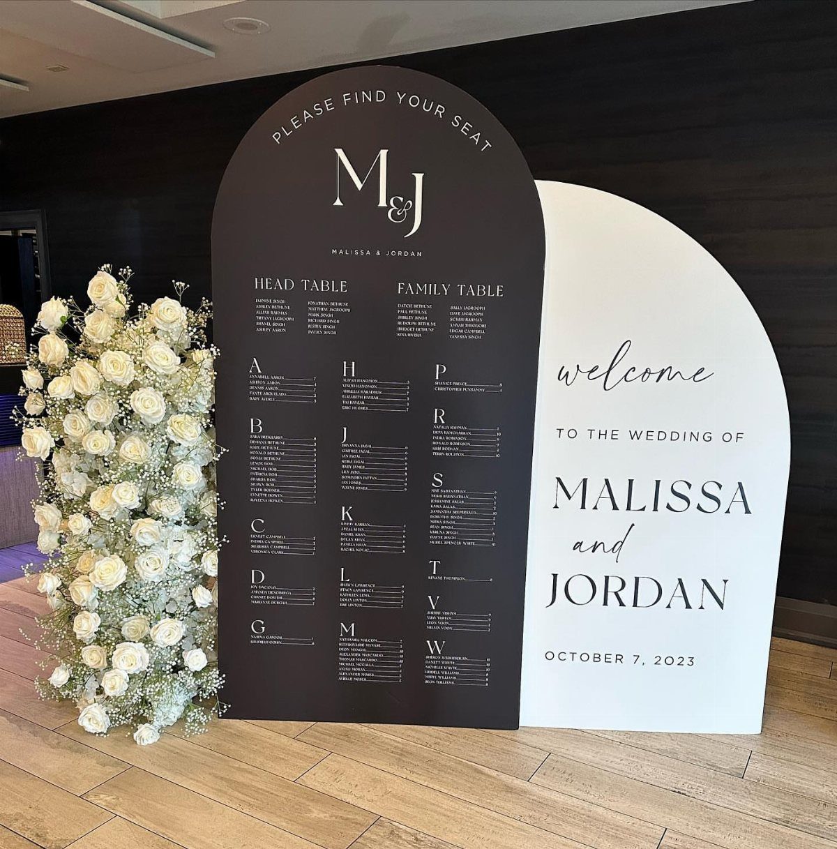 Black arched alphabetical seating chart and white welcome sign with white floral arrangement.