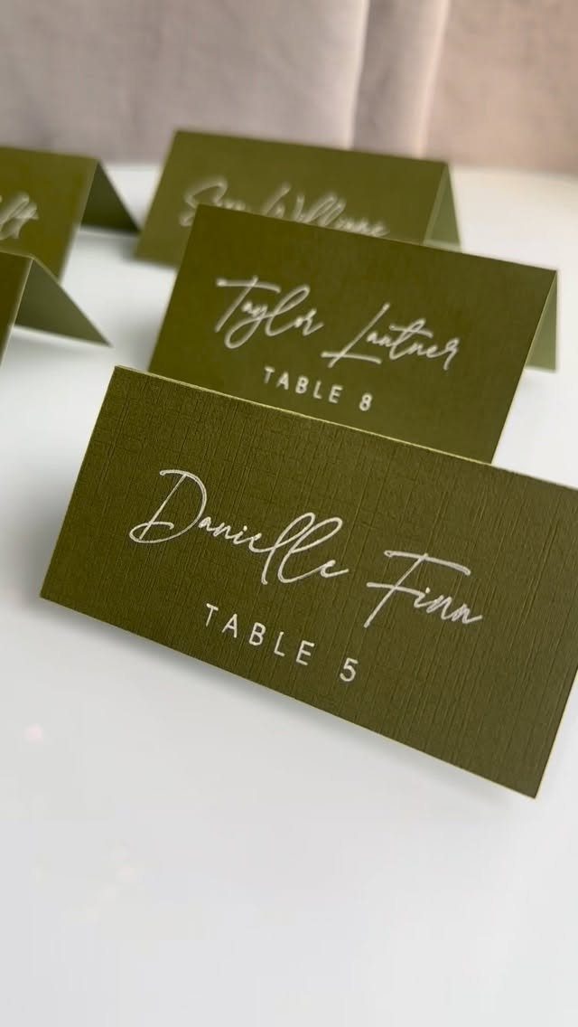 Olive green place cards with white cursive names and table numbers on white surface