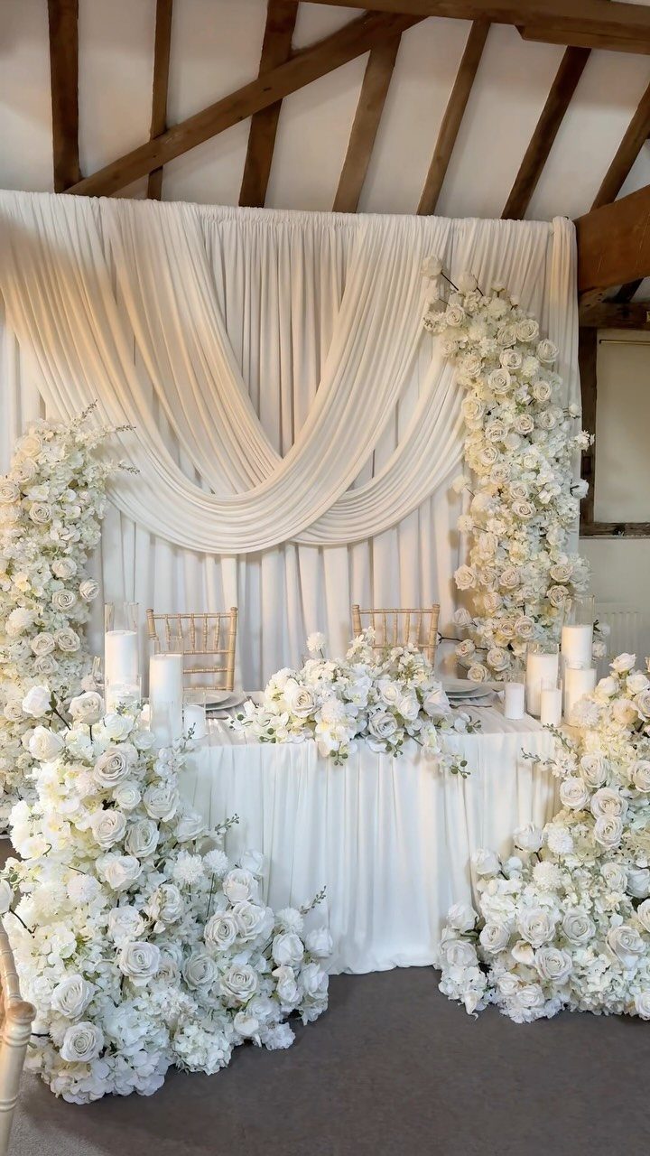 Small white sweetheart table with floral arrangements and a draped fabric backdrop.