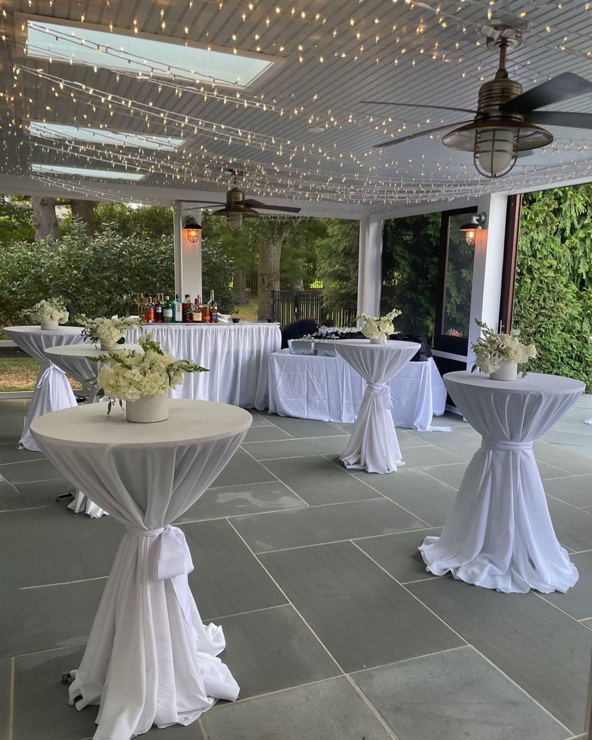 High-top cocktail tables with white linens and flowers on a tiled outdoor patio.