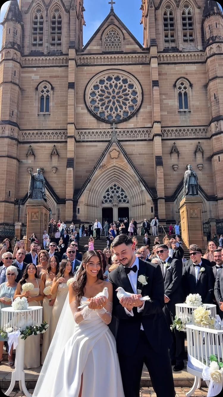 Bride and groom holding white doves in front of a stone cathedral with guests.