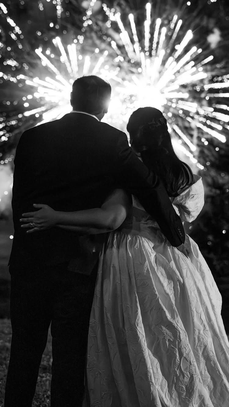 Newlywed couple embracing while watching a large firework burst in the night sky.
