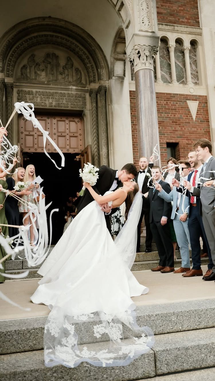 Bride and groom kissing on church steps while guests wave white ribbon wands.