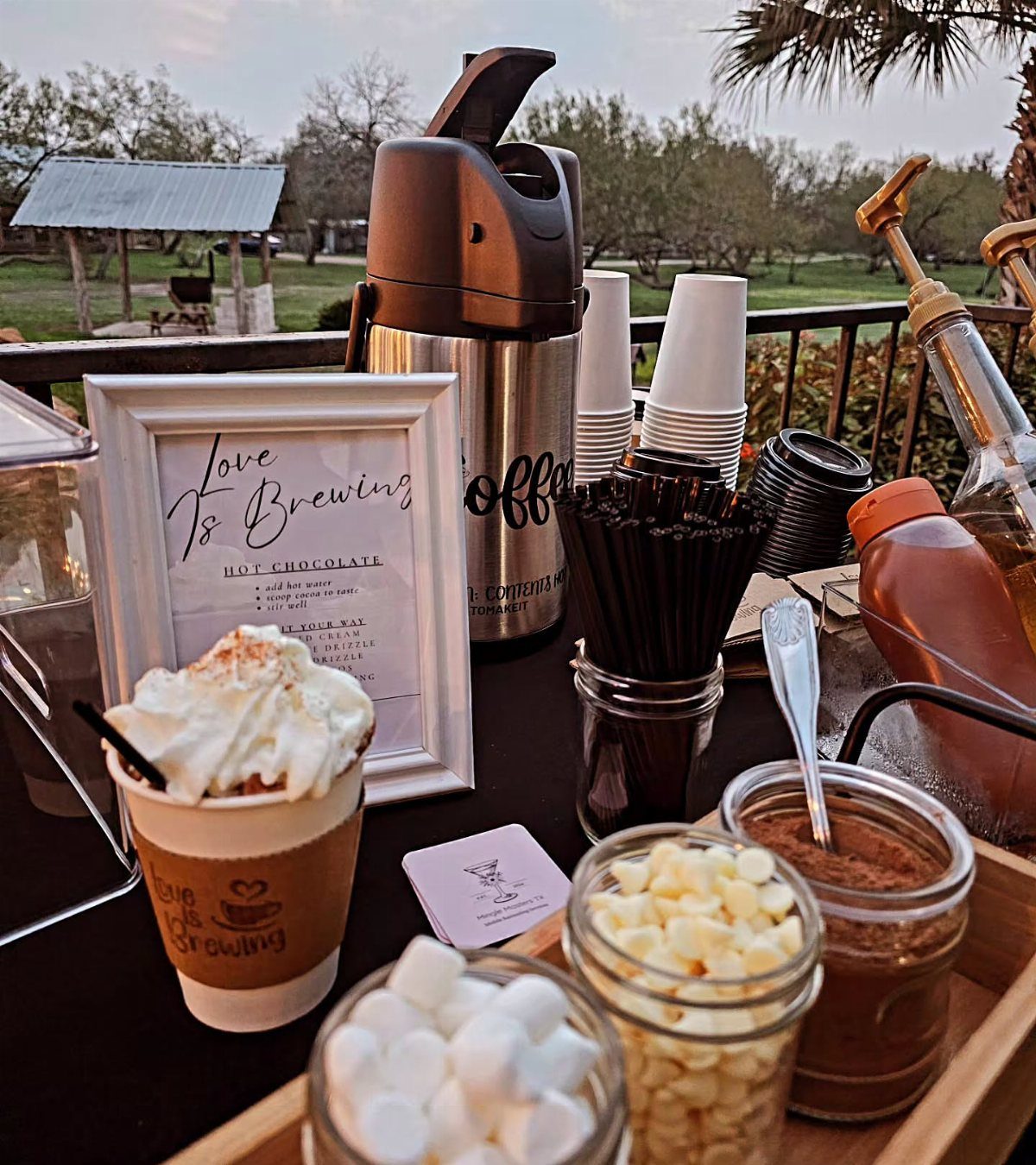 Hot cocoa bar with thermal dispenser, marshmallows, white chocolate, and Love is Brewing sign.