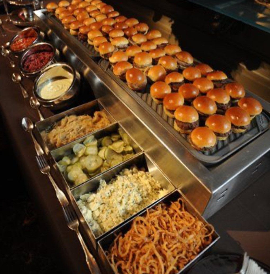 Mini sliders on a buffet tray with various toppings in metal containers.
