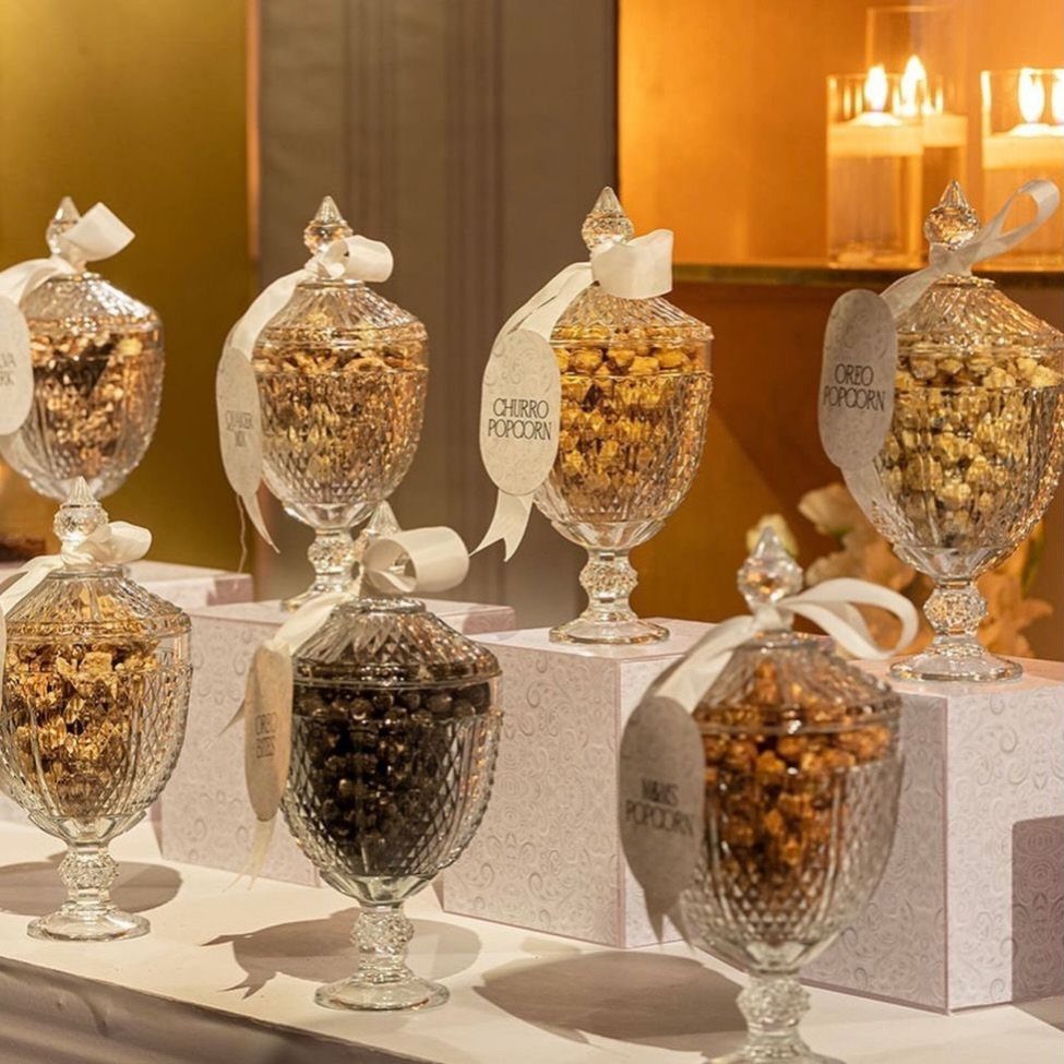 Glass apothecary jars filled with flavored popcorn on white pedestals with white ribbons.