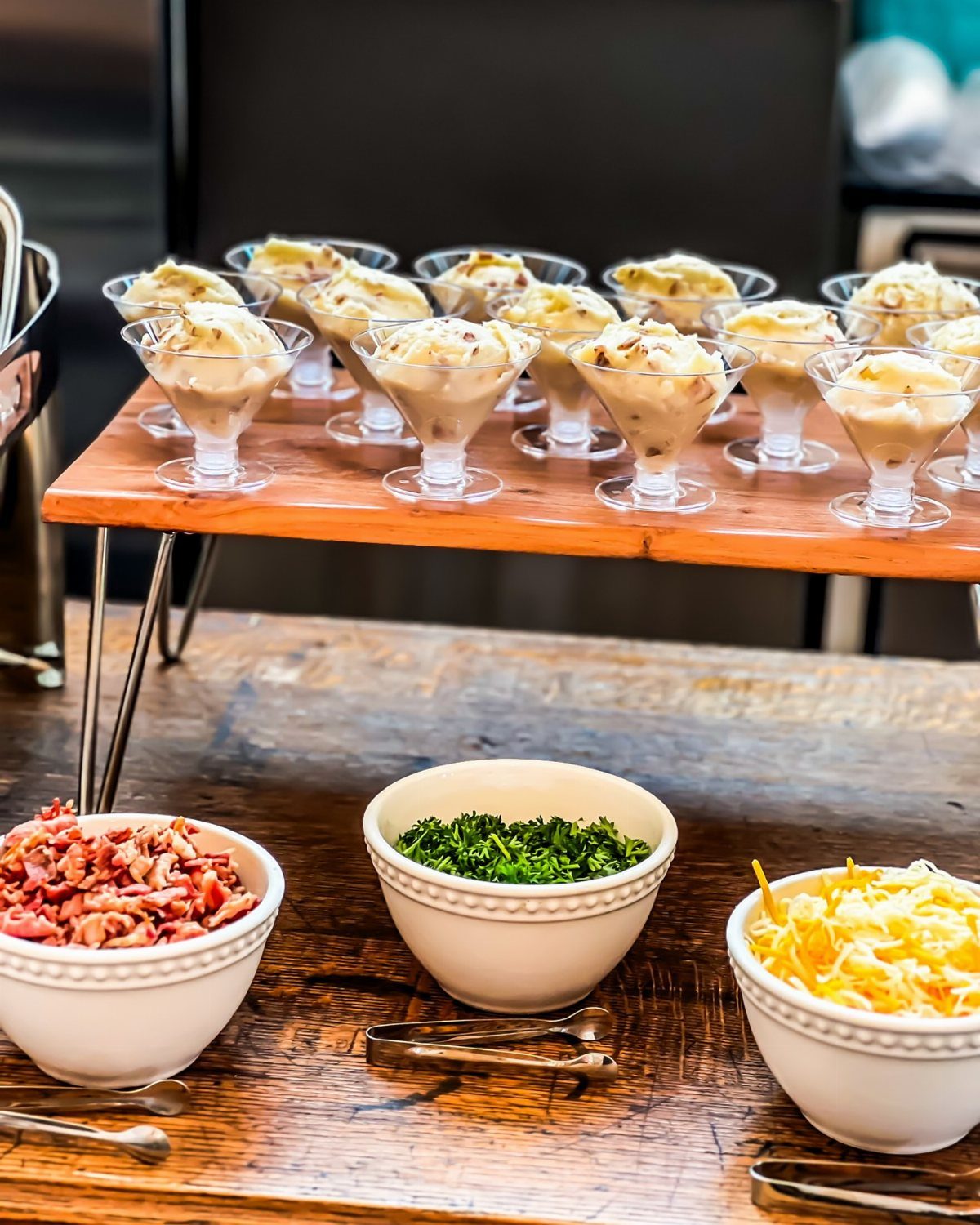 Mashed potato bar with toppings like bacon, chives, and cheese on wooden tables.