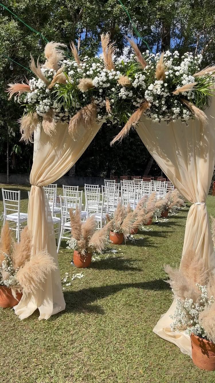 Wedding arch with cream drapery, white flowers, and pampas grass on green grass.