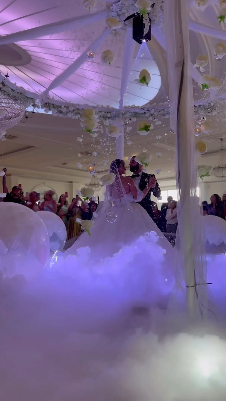 Bride and groom dancing under a white floral dome with purple lighting and fog.