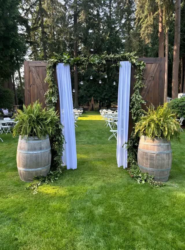 Wooden wedding altar with white drapes, greenery, and wine barrels on green grass.