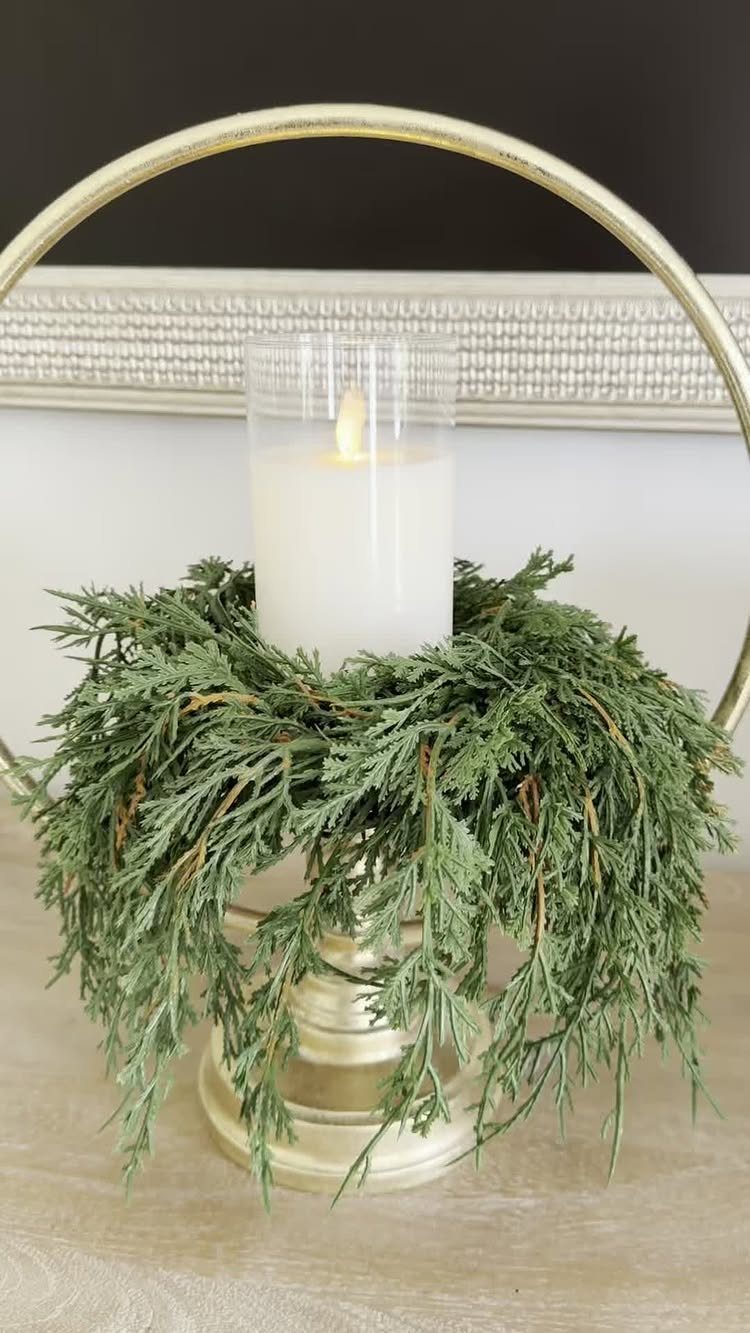 White pillar candle surrounded by green cedar foliage in a gold hoop frame.