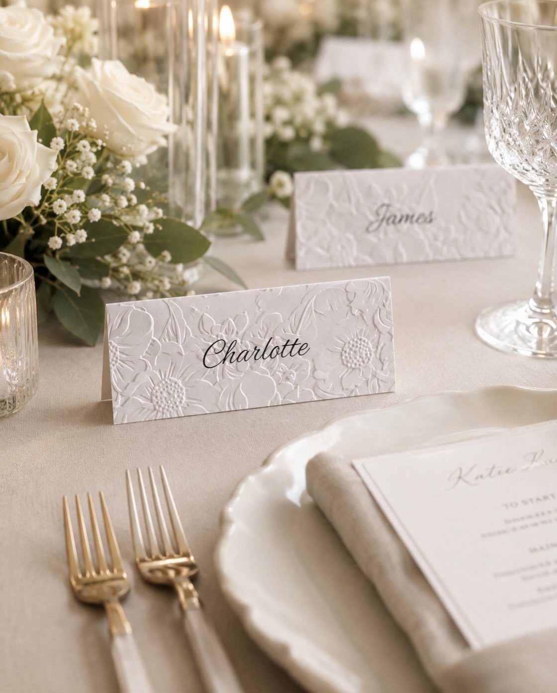 Embossed white place cards and gold cutlery on a wedding table with white roses.