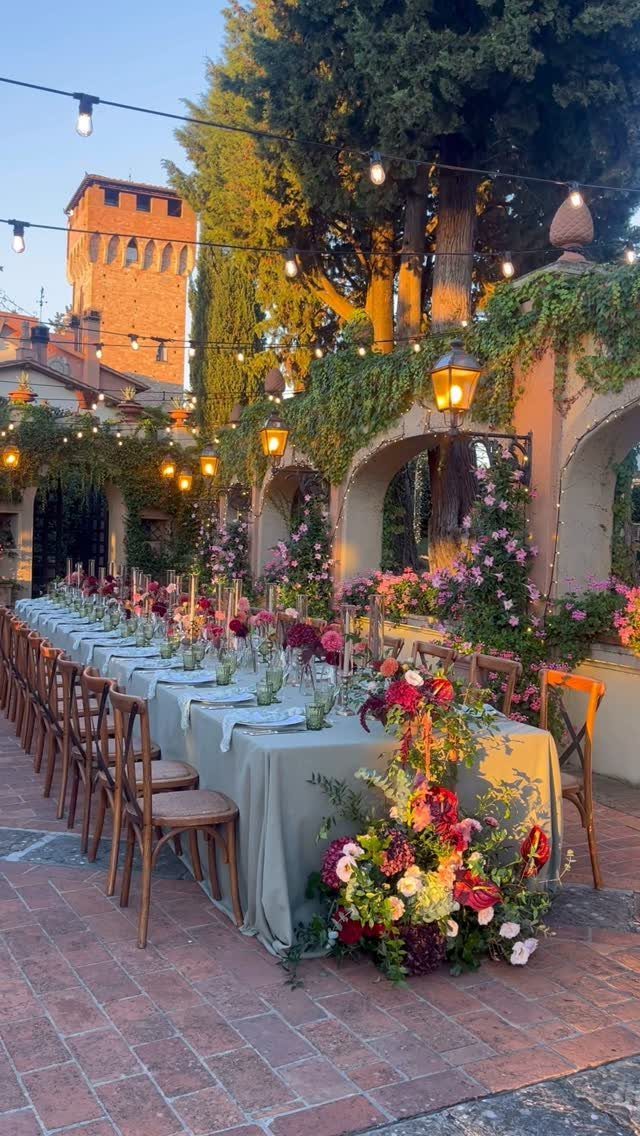 Long wedding table with vibrant red and yellow floral arrangements under outdoor string lights.