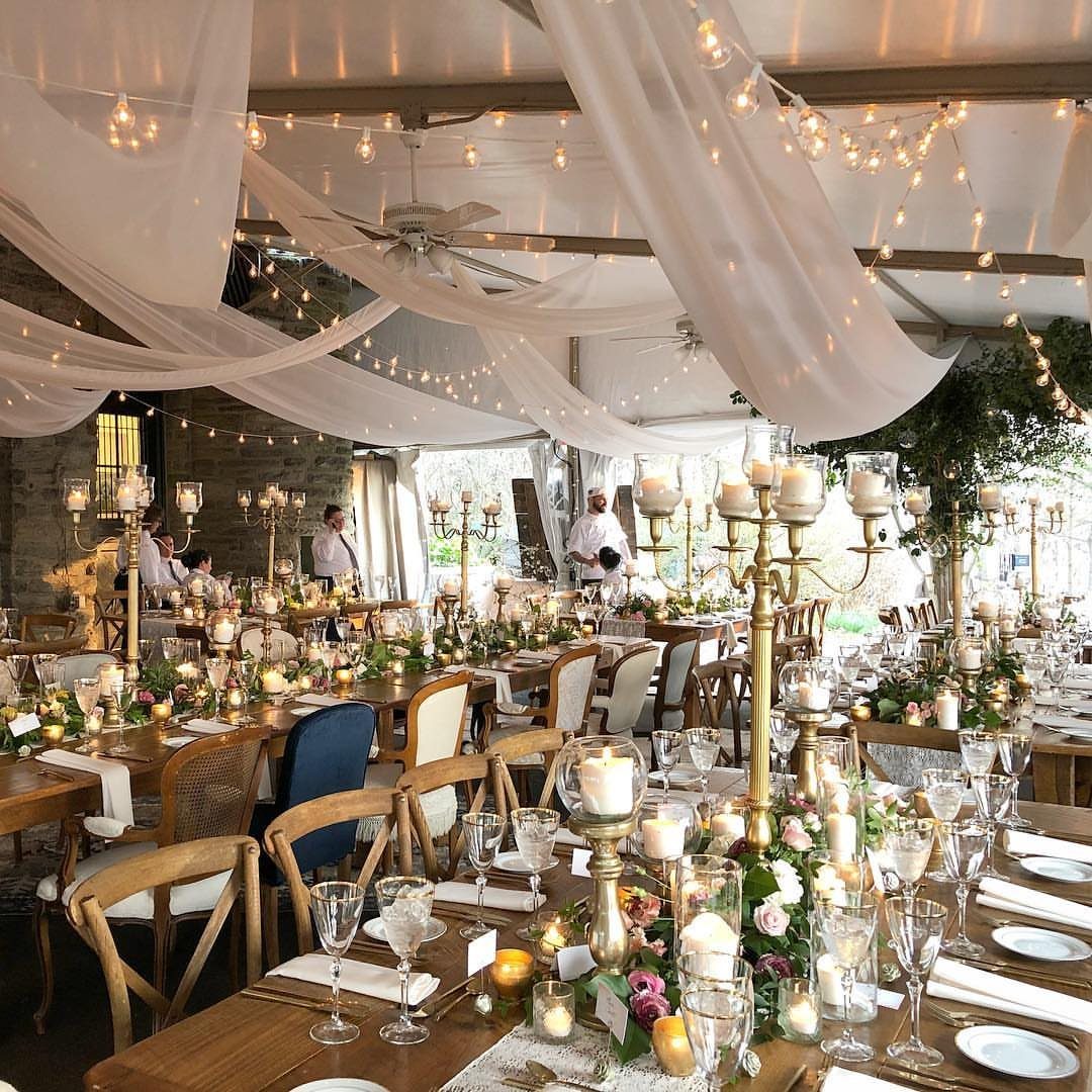 Wedding reception under a white tent with draped fabric, fairy lights, and gold candelabras.