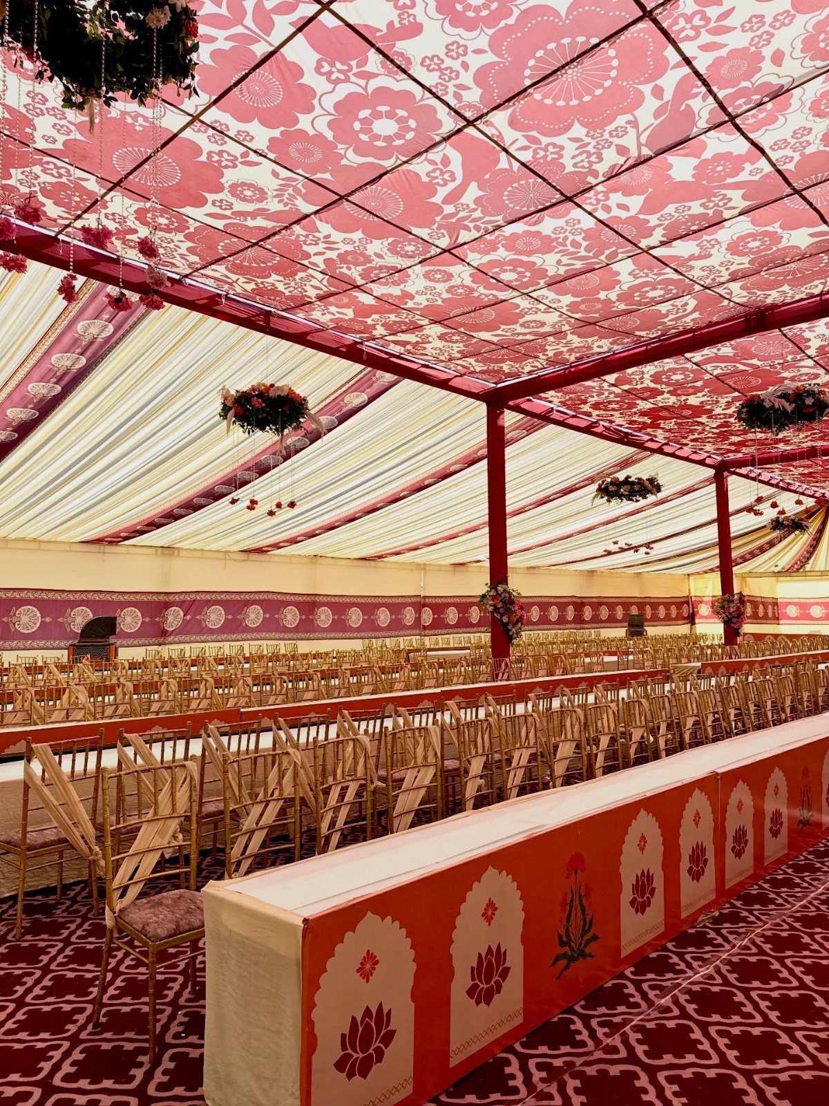 Decorated wedding tent with red patterned ceiling, gold chairs, and long banquet tables.