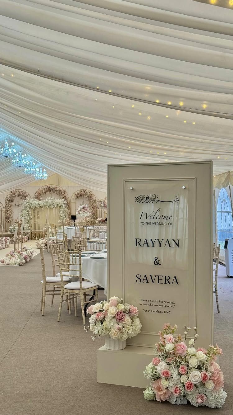 Wedding welcome sign with floral arrangements inside a white draped tent with gold chairs.