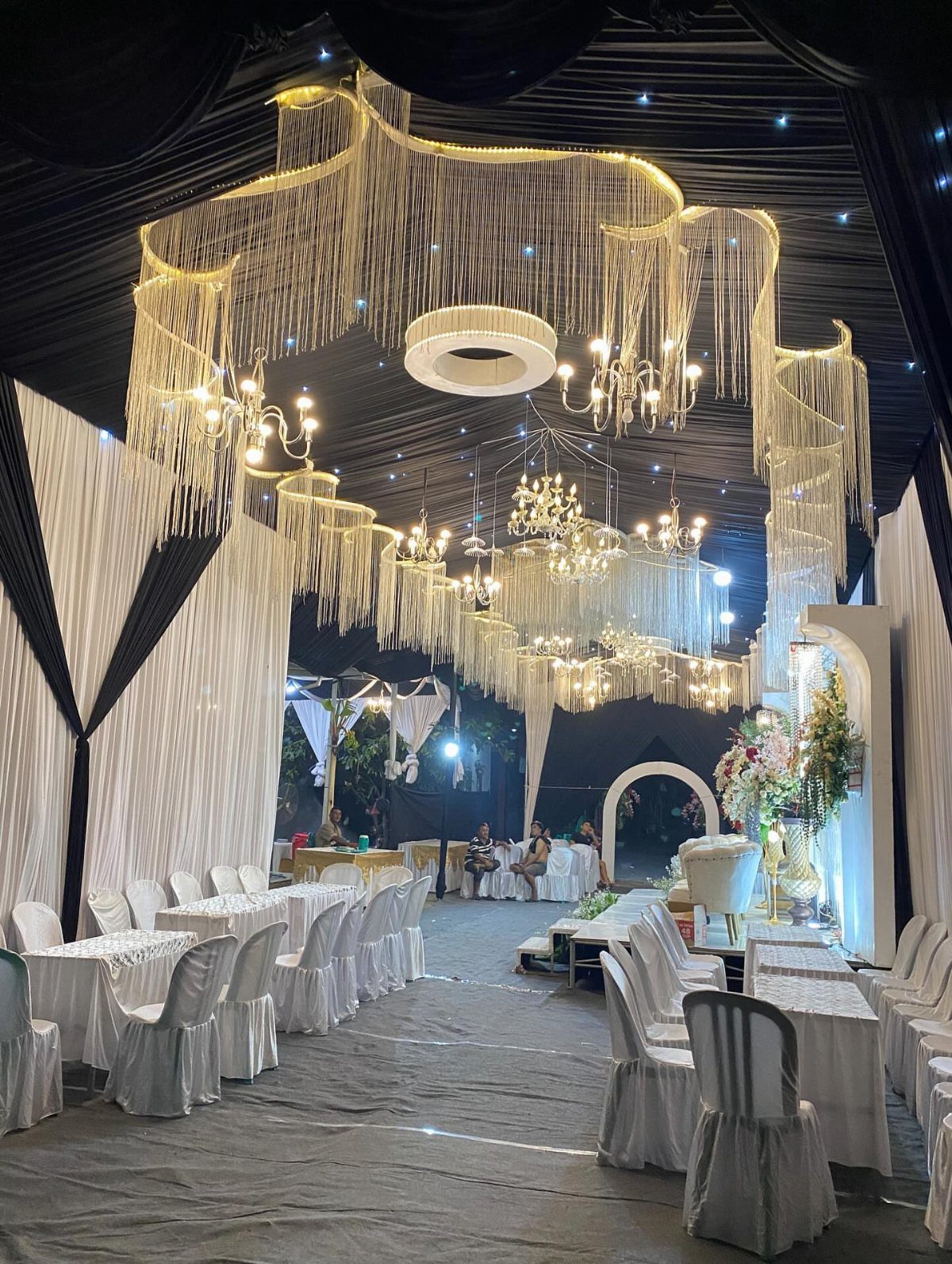 Wedding tent interior with golden shimmering ceiling decorations, chandeliers, and white banquet tables.