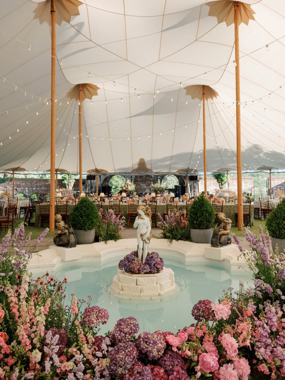Circular stone fountain with a statue surrounded by pink and purple flowers inside a tent