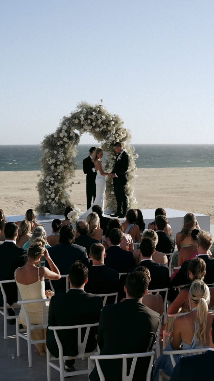 Bride and groom under a floral arch on a beach with formal guests.
