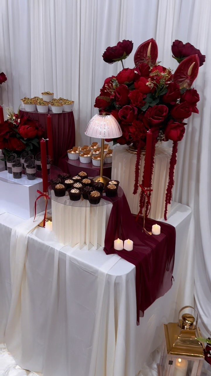 Wedding dessert table with burgundy runners, red roses, and gold accents on white linens.