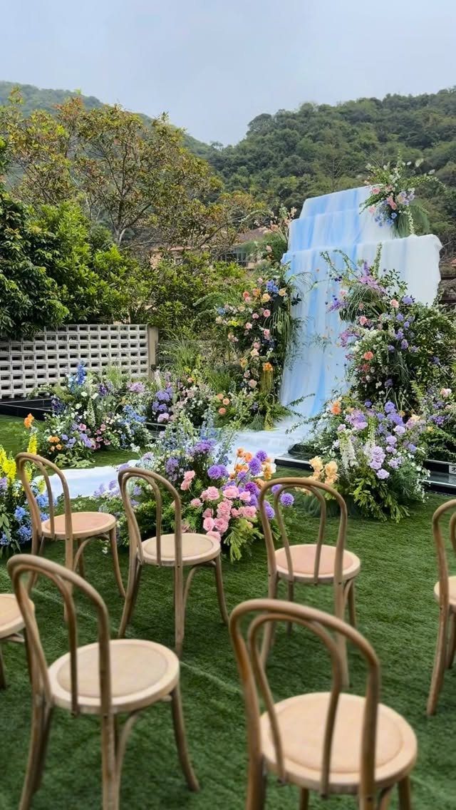Outdoor wedding ceremony setup with light wood chairs and colorful floral arrangements.