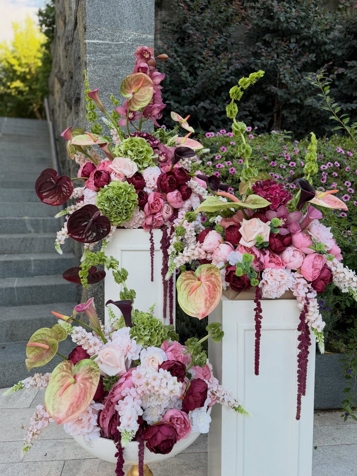 Burgundy and purple floral arrangements on white pedestals against a stone wall.