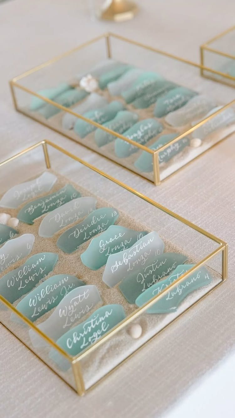 Sea glass place cards in gold glass boxes resting on white sand.
