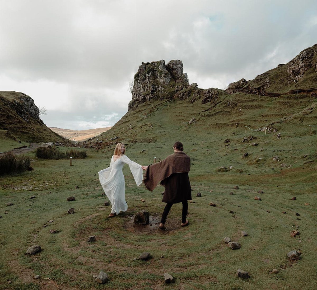 Couple in rustic attire holding hands in a green valley with rocky cliffs.