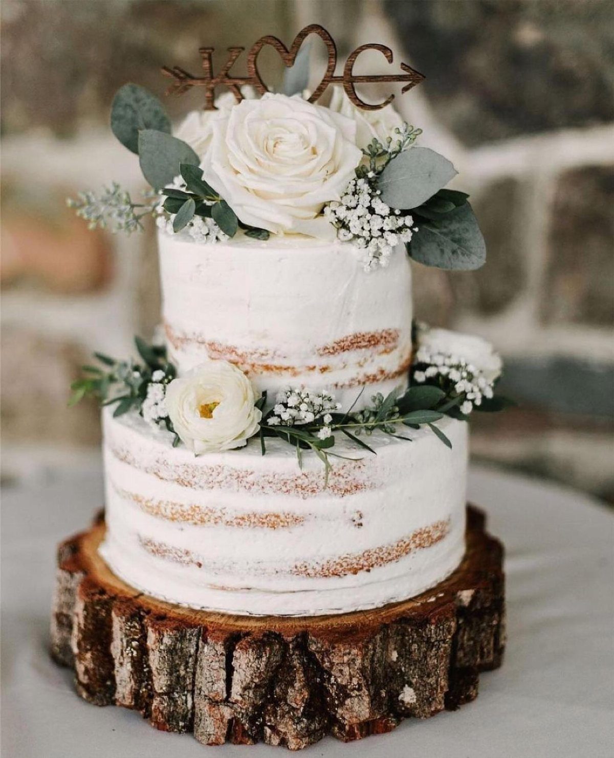 Two-tier semi-naked wedding cake decorated with white roses and greenery on a wood slice.