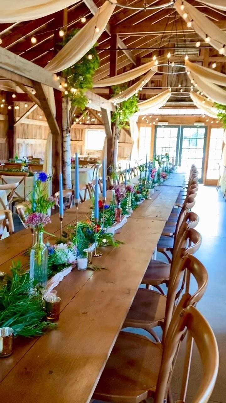 Long wooden wedding table with greenery, wildflowers, and blue candles in a barn.