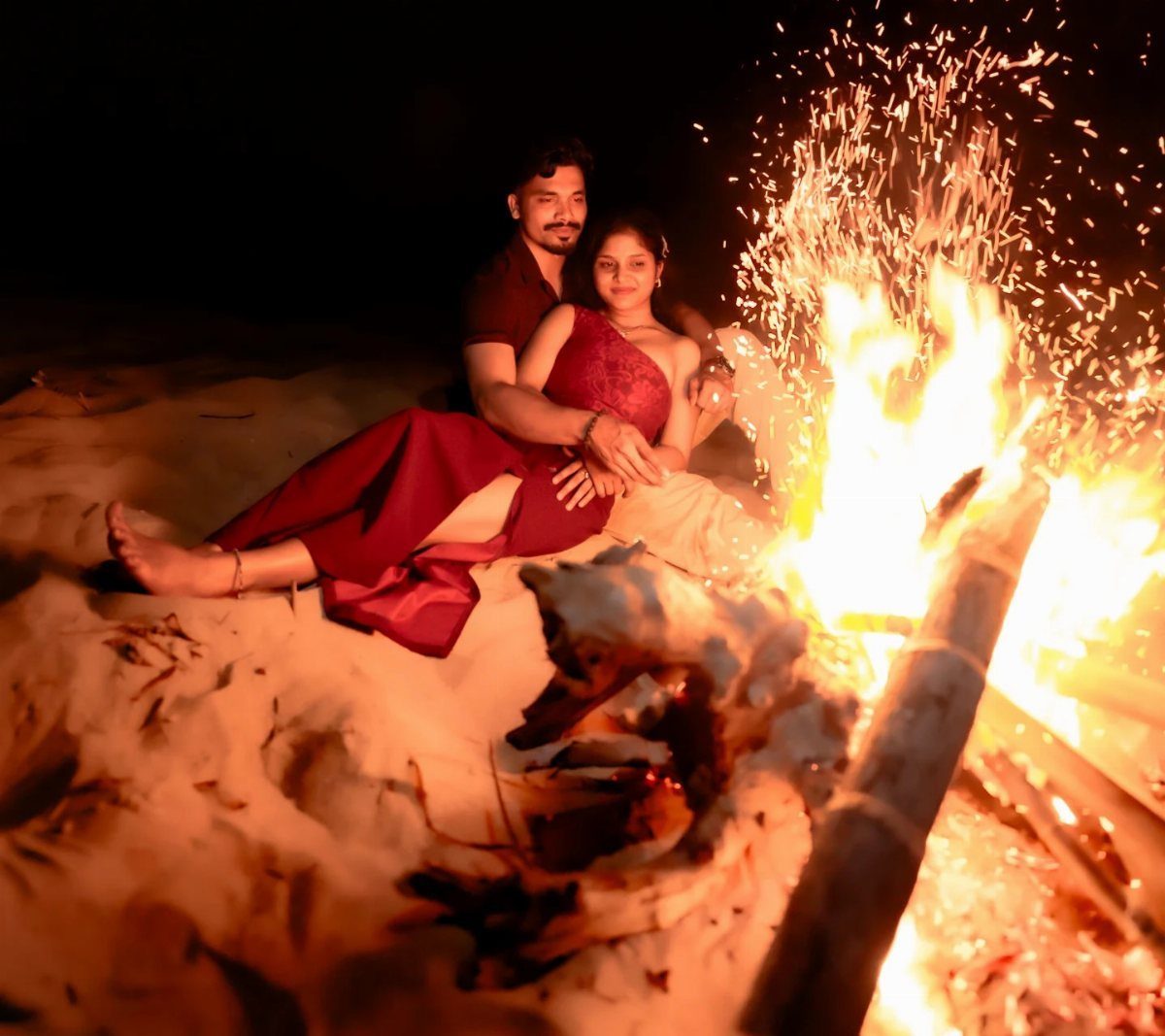 Couple in red clothing sitting on sand beside a large beach bonfire.