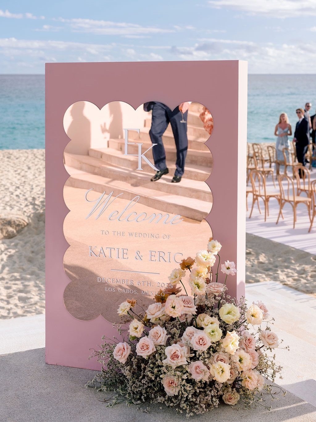 Pink wedding welcome sign with floral arrangement on a sunny beach shoreline
