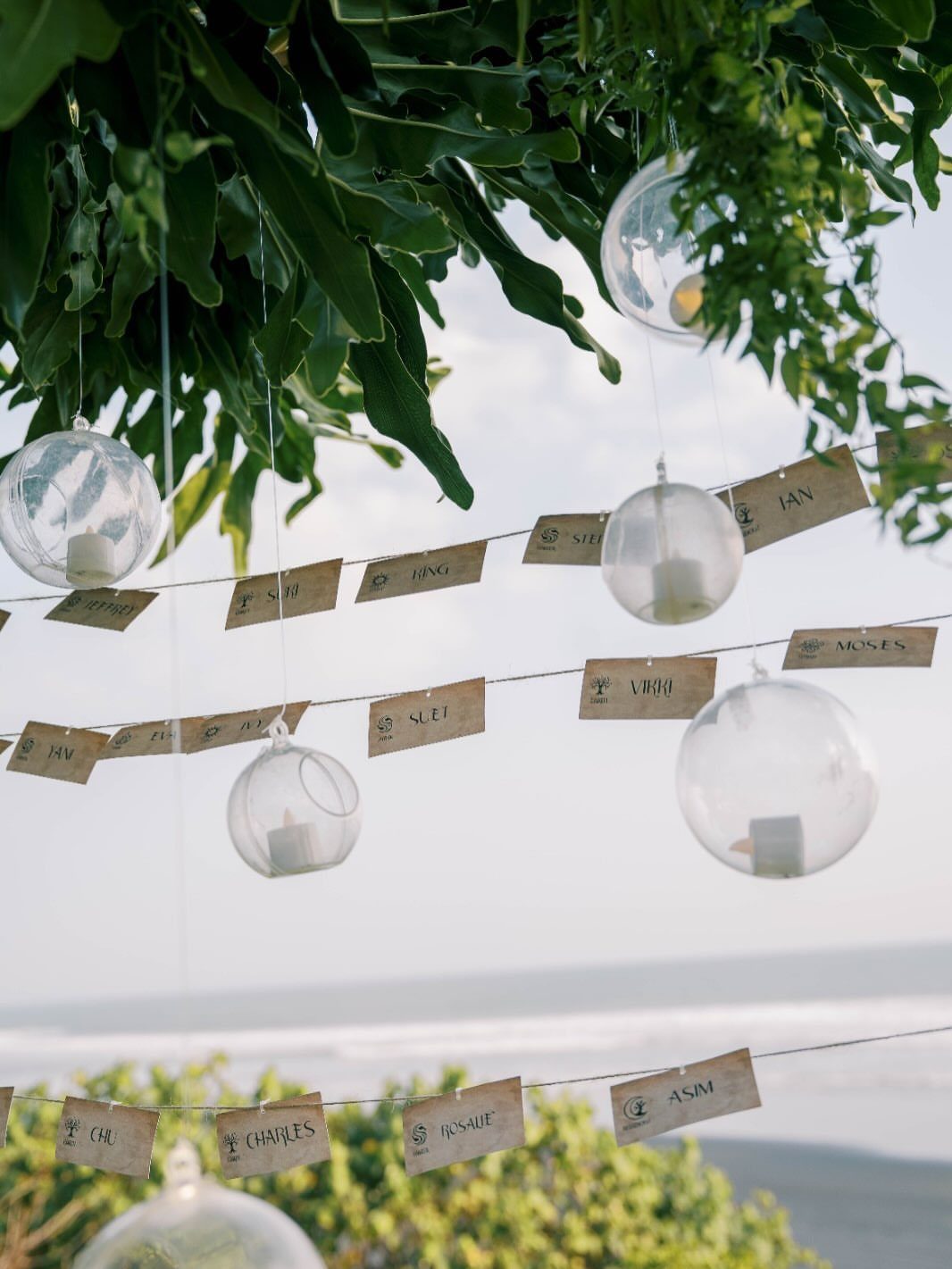 Glass globe candles and wooden name tags hanging from tropical green leaves by beach
