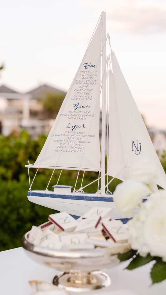 Model sailboat with a drink menu printed on the sail near white flowers