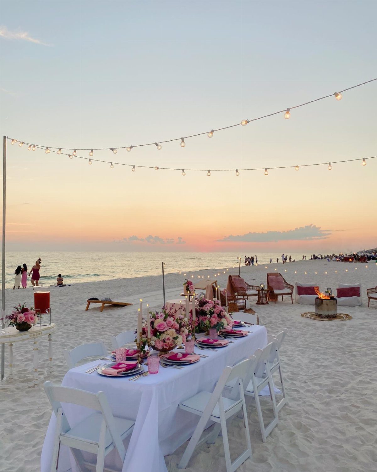 Elegant beach dinner table with pink flowers and string lights at sunset.