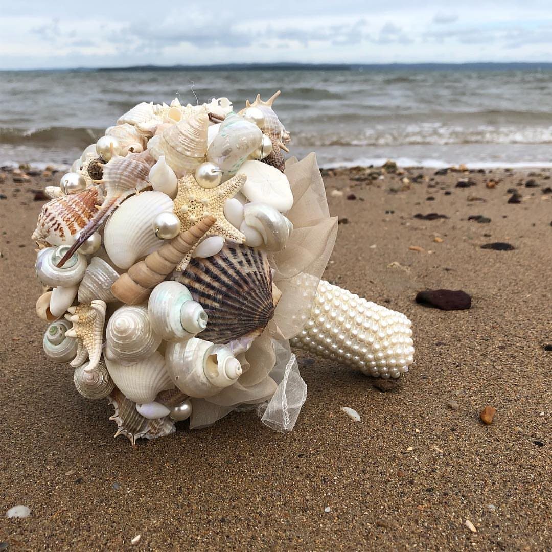 Seashell bridal bouquet with pearls resting on a sandy beach by the ocean.
