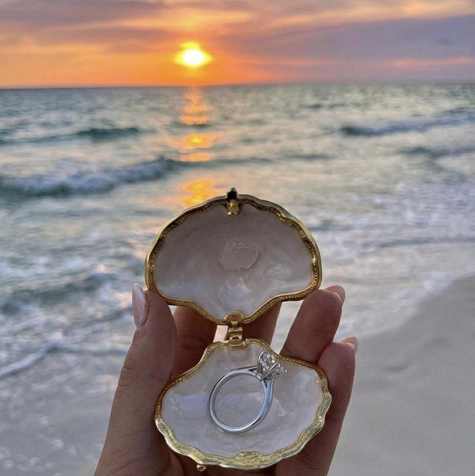 Diamond engagement ring inside a gold-edged seashell on a beach at sunset.