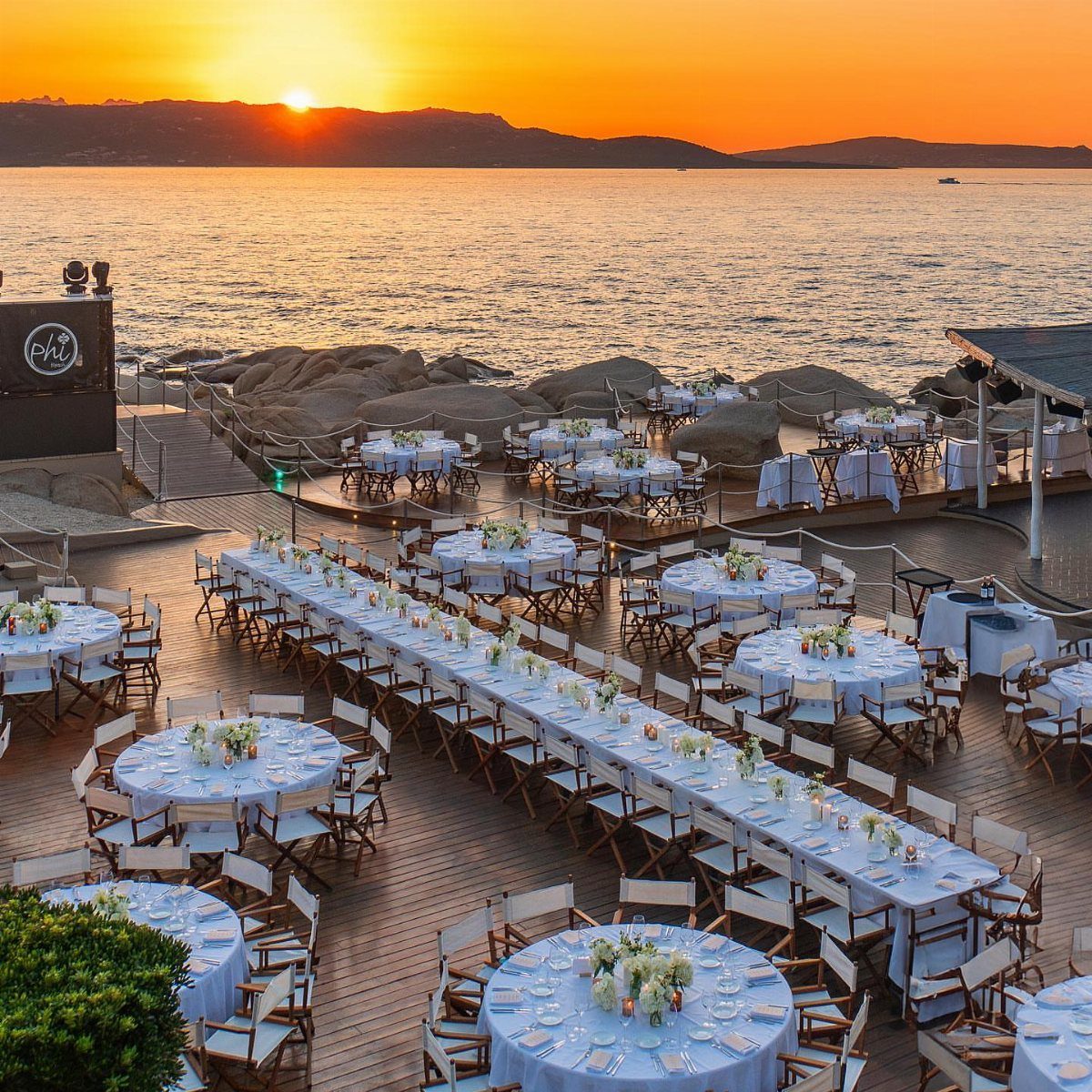 Outdoor seaside dining setup with white tables and chairs during a golden sunset.
