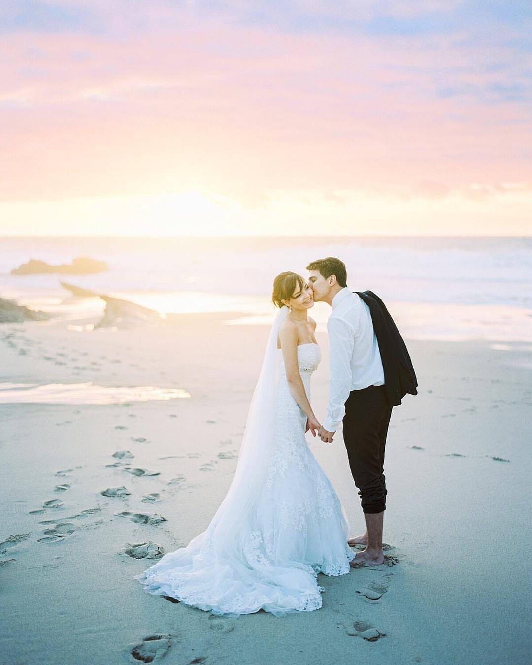 Barefoot bride and groom kissing on a sandy beach during a pink sunset.