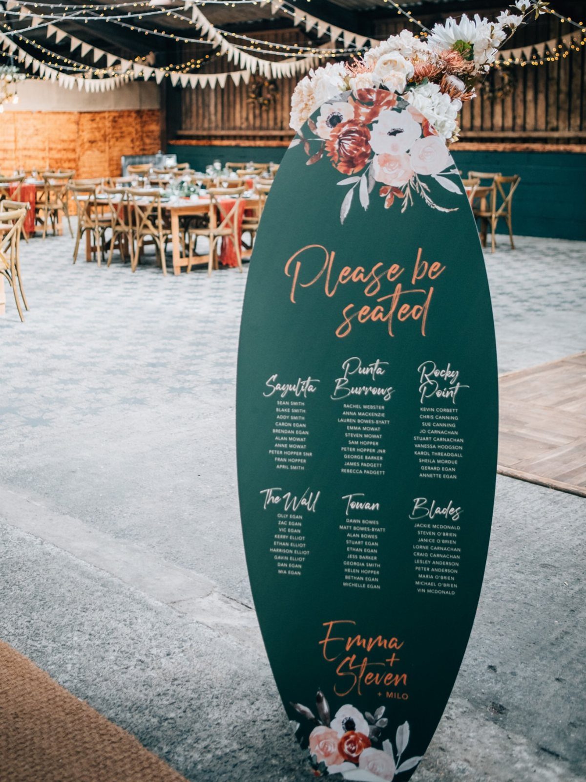 Dark green surfboard seating chart with gold text and floral accents at a wedding.