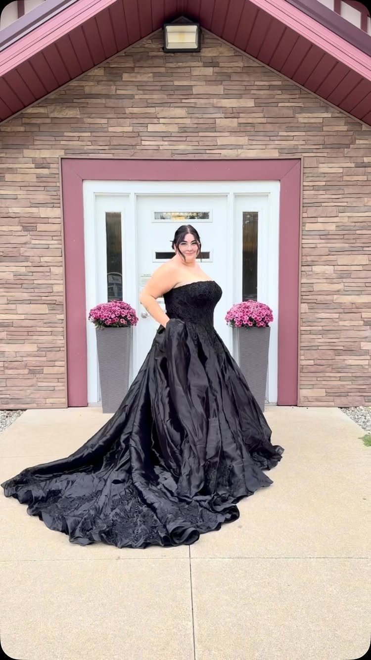Woman in a strapless black ballgown wedding dress posing before a stone house.