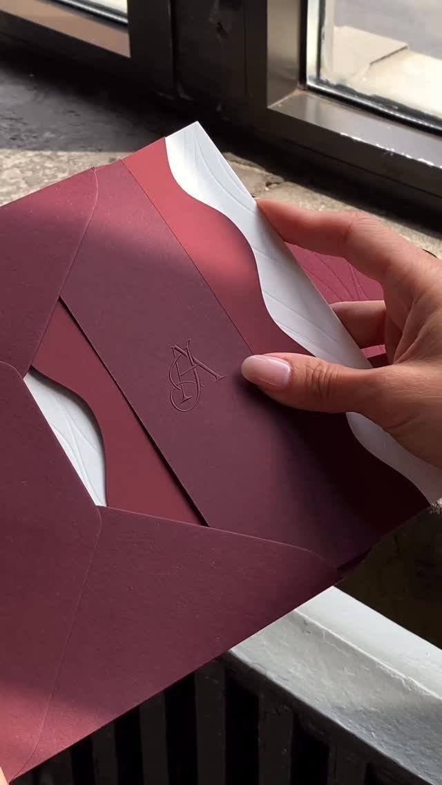 Burgundy wedding invitation suite with a white wavy paper insert held by a hand.