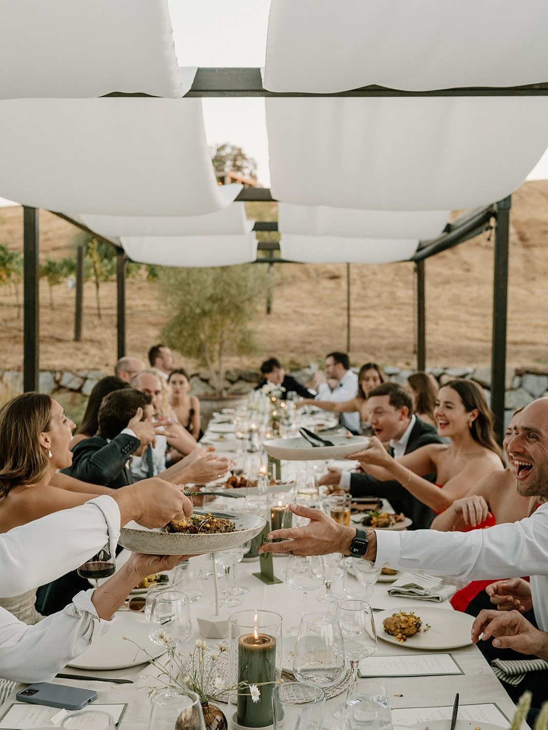 Guests at a long outdoor wedding table passing platters of food and laughing.