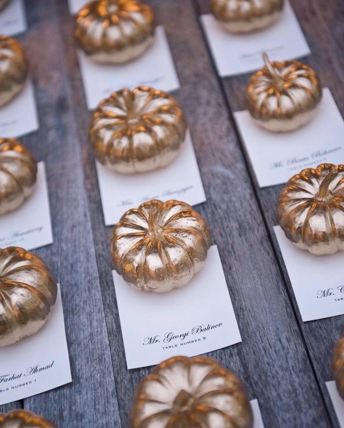 Gold painted mini pumpkins on white name cards atop a grey wooden table.