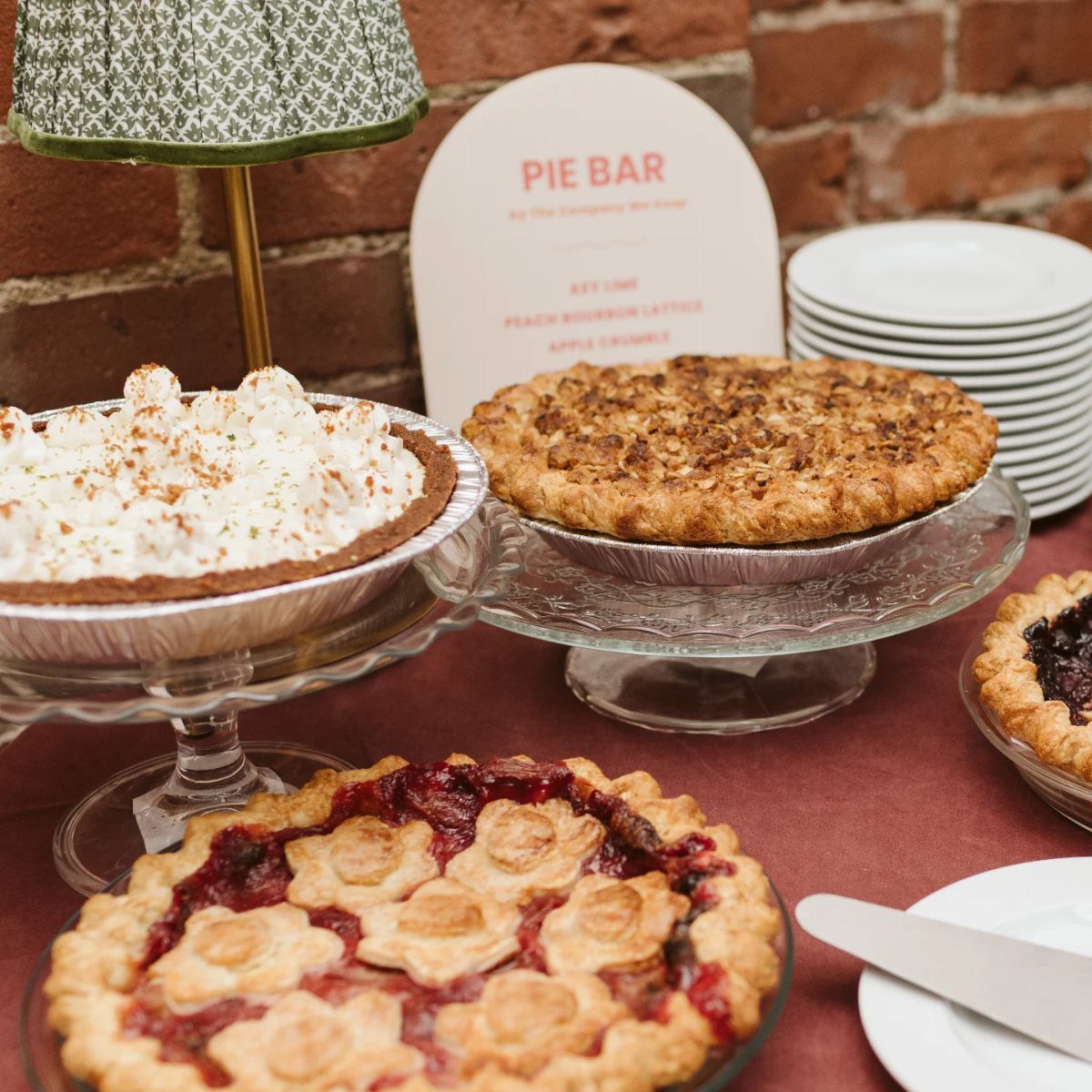 Assorted pies on glass stands with a Pie Bar sign on red tablecloth.