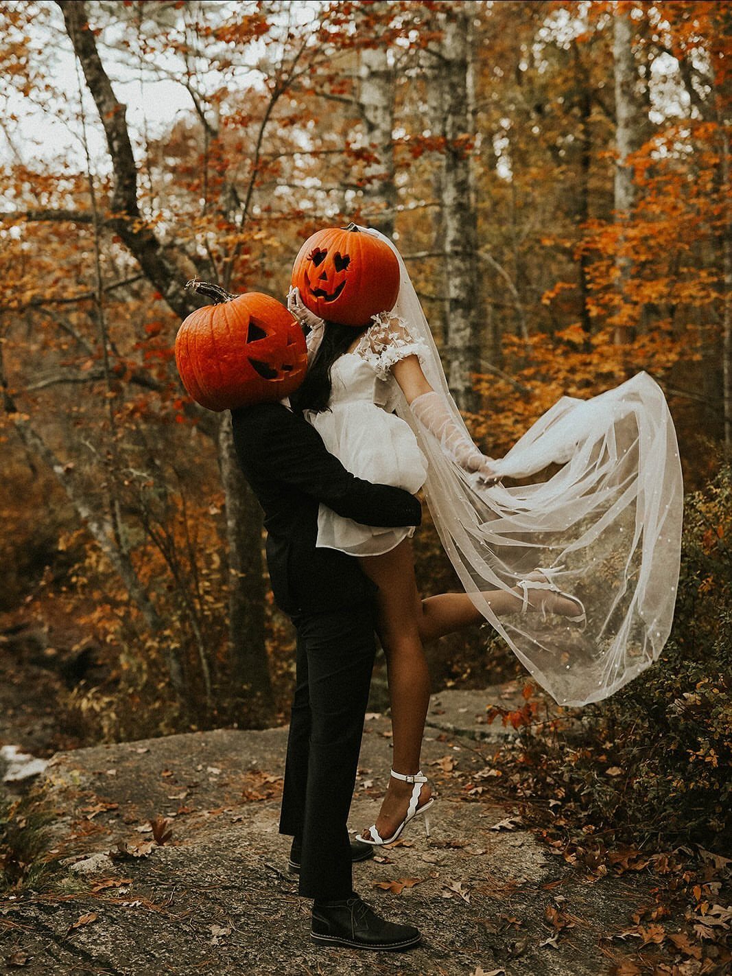 Couple in wedding attire wearing carved pumpkin heads in an autumn forest.