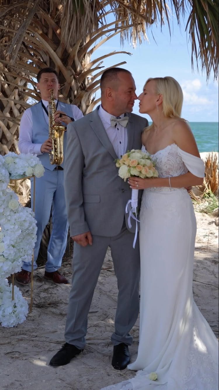 Bride and groom kissing on a sandy beach with a saxophone player behind them.