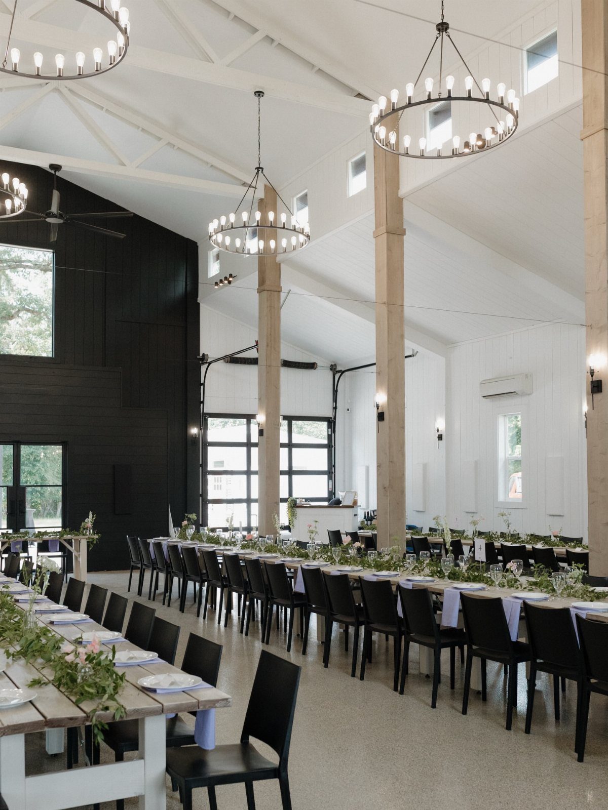 Modern farmhouse wedding reception with long wooden tables, black chairs, and circular chandeliers.