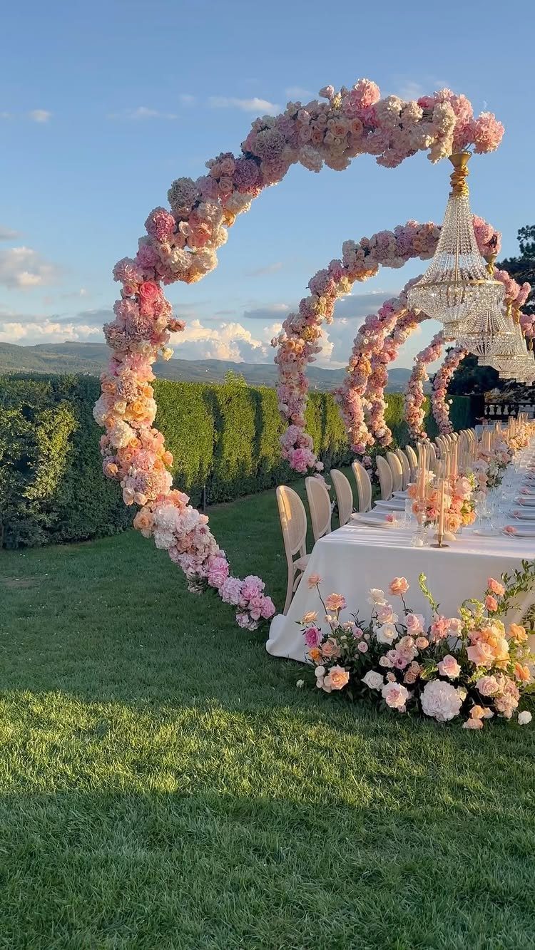 Crystal chandelier hanging from pink floral arches over a wedding reception table outdoors.